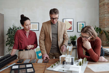 Young architects surrounding mature male colleague in smart casualwear while choosing parts for...