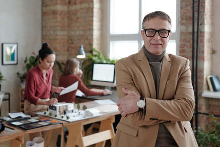 Smiling mature male architect in smart casualwear and eyeglasses standing against his two young...