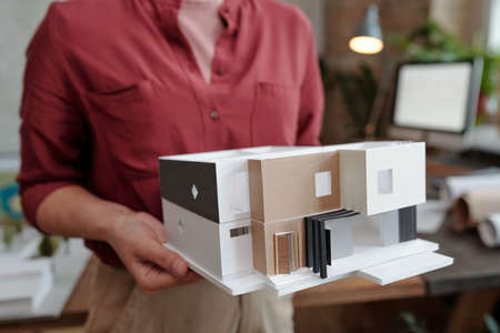 Hand of young female architect or engineering designer in casualwear holding paper model of new house while standing in officeの写真素材