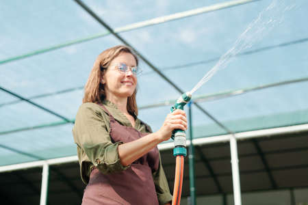 Cheerful brunette female gardener in eyeglasses and brown apron watering plants in hothouse while standing in front of cameraの写真素材