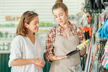 Happy young assistant of gardening shop in apron and shirt showing small shovels to mature brunette female customer in large supermarketの写真素材