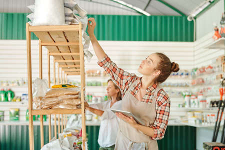 Pretty assistant in workwear standing by shelf with packed fertilizers and checking bar codes on...