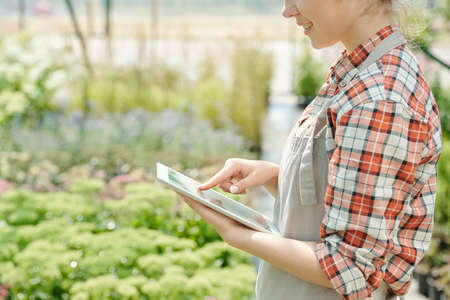 Side view of happy young female worker of greenhouse pointing at display of touchpad while scrolling through images of garden plantsの写真素材
