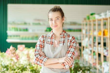 Young pretty female farmer or shop assistant with crossed arms standing in front of camera and...