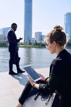 Young businesswoman in formalwear and eyeglasses using digital tablet while sitting by riverside in front of African businessman textingの写真素材