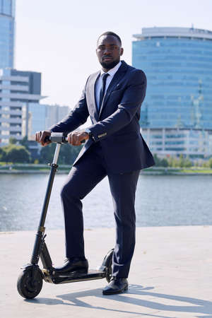 Young serious African businessman in formalwear standing on electric scooter by riverside against...