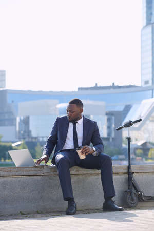 Young African businessman with laptop and coffee preparing presentation while sitting by riverside against skyscrapers and modern buildingsの写真素材