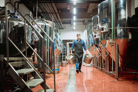 Young male worker of contemporary brewery in workwear and protective mask carrying two cisterns...