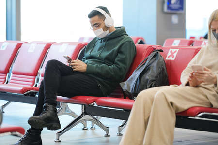 Young man with headphones listening to music and scrolling in smartphone while sitting in lobby of modern airport and waiting for flightの写真素材