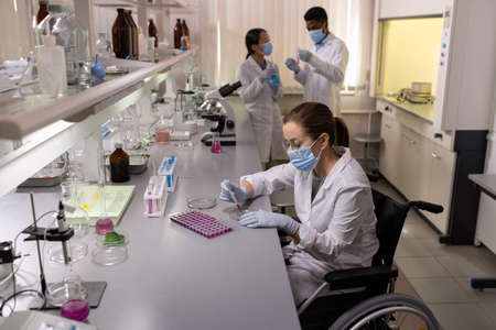 Chemist in mask examining liquid in test tubes while working with her colleagues in the laboratory