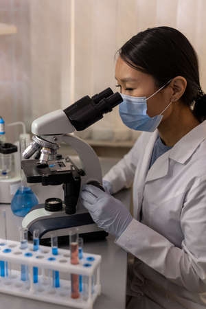 Asian young biologist in mask looking through the microscope and examining samples in the labの写真素材
