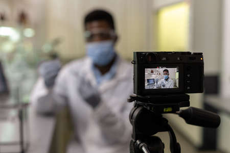 African young scientist holding test tube with liquid and looking at camera he recording a video for...