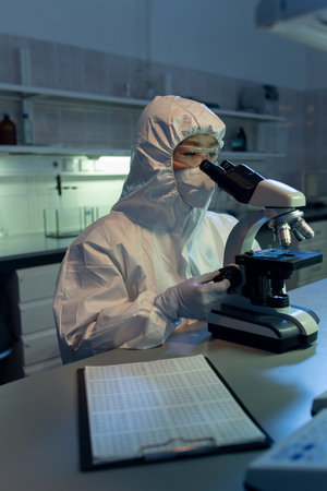 Female chemist in protective wear sitting at her workplace and looking through the microscope while working in the labの写真素材