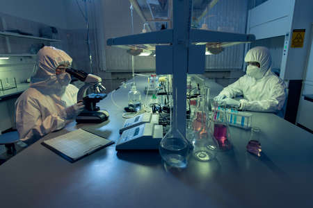 Two colleagues in protective workwear sitting at the table and working with chemical samples in test tubes in the laoratoryの写真素材
