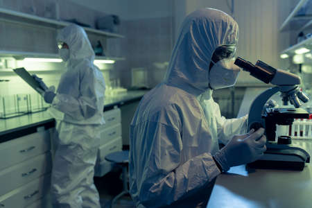 People in protective workwear working in the laboratory and examining samples through the microscopeの写真素材