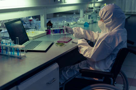 Disabled chemist in protective workwear and mask examining liquid in test tubes at the table in the...