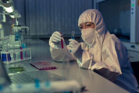 Female chemist in protective workwear sitting at the table and holding test tubes with samples and...