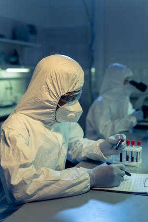 Scientist in protective workwear working at the table with danger chemical samples with his...