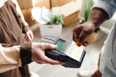Hand of young man in workwear holding payment terminal while African male paying for loading and...