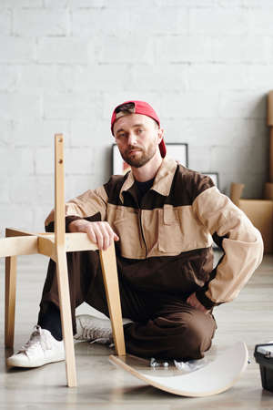 Serious young furniture assemblage specialist in workwear sitting on the floor by unfinished chair...