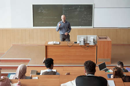 Bald mature professor in smart casualwear standing by desk in front of audience and explaining subject of lecture to intercultural studentsの写真素材