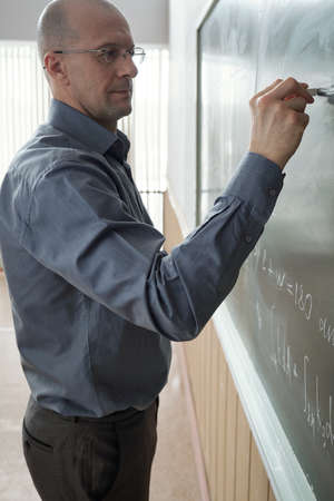 Mature serious professor of university standing in front of blackboard in auditorium or lecture...