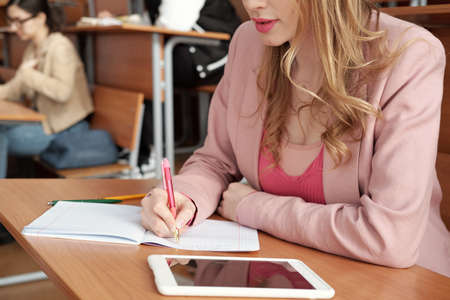 Girl with long blond hair making notes in copybook at lecture while sitting by desk against...