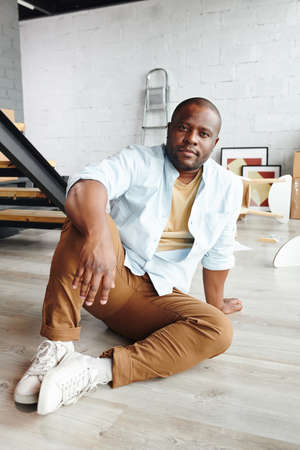 Young African man in brown pants, blue shirt and white cross-shoes sitting on the floor in front of...