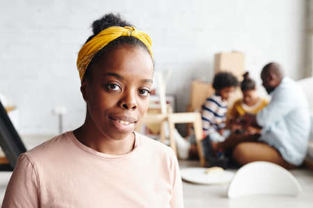 Happy African woman in yellow headband and beige t-shirt standing against her husband and their two children assembling furnitureの写真素材