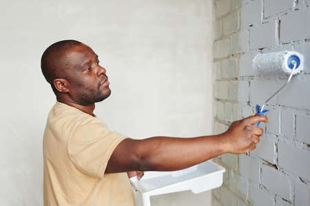 Young African man in beige t-shirt holding paintroller while standing in front of brick wall of living-room and painting it into white colorの写真素材