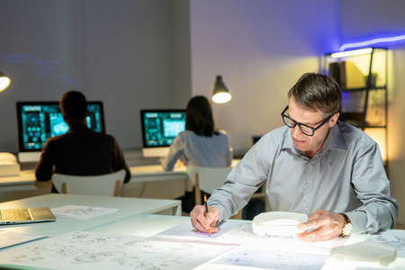 Concentrated mature automotive designer in eyeglasses sitting with car model at table and editing...