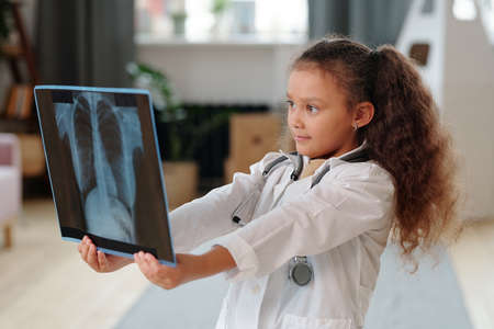 Little girl in white coat holding x-ray image and examining it she playing in hospital at homeの写真素材