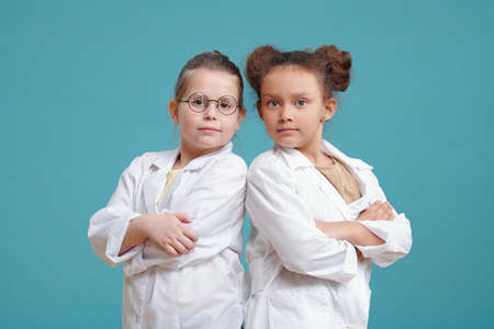 Portrait of two little girls in white coats standing with arms crossed and looking at camera against...