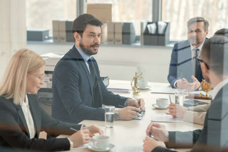Confident young bearded lawyer in suit sitting at conference table and discussing contract content with colleagues, behind glass viewの写真素材