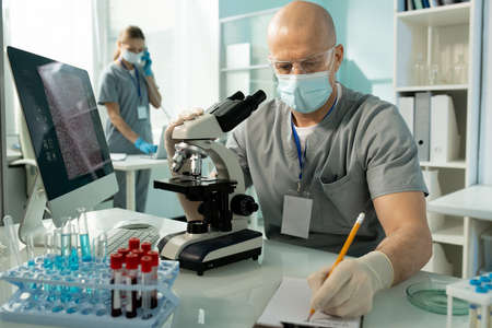 Busy lab worker in mask and gloves sitting at table and making notes about samples while using...