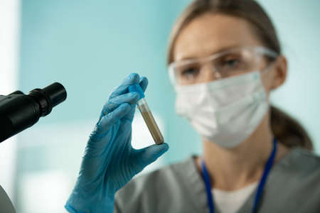 Close-up of young laboratory worker in mask and blue glove holding test tube of muddy liquid while working with samples and microscopeの写真素材