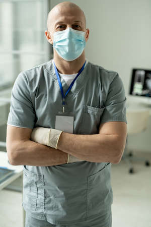 Portrait of satisfied bald surgeon in facial mask and gloves standing with crossed arms in modern...