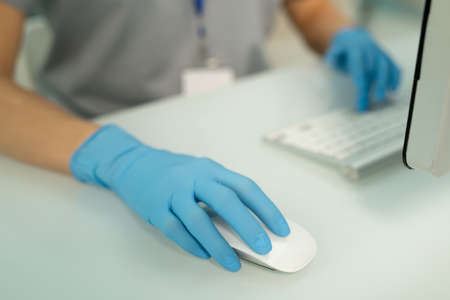 Close-up of unrecognizable nurse in blue gloves sitting at desk and using computer mouse while...