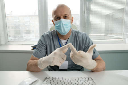 Portrait of bald middle-aged doctor in mask and gloves sitting at table and counting fingers while...