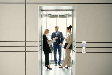 Serious business people standing in elevator and preparing for meeting or surfing net on gadgets