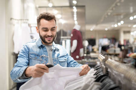 Happy young man with shirt in hands choosing new clothes in storeの写真素材