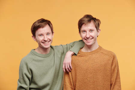 Portrait of happy young man in casual sweater leaning on shoulder of twin brother against orange backgroundの写真素材