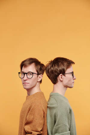 Portrait of serious handsome young man in glasses standing back to back with twin brother against...