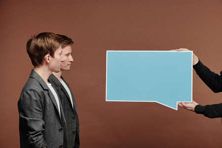 Side view of serious young twin brothers in suits standing in line and looking at blue speech tag