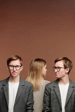 Rear view of blond-haired woman standing behind two jealous twins in eyeglasses, brown background
