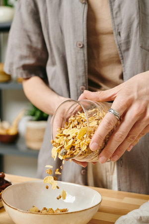 A person in casualwear putting corn flakes into a bowl while preparing breakfastの写真素材