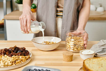 A woman pouring milk into bowl with corn flakes by kitchen tableの写真素材