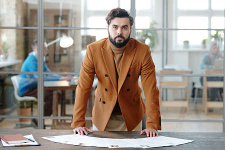 Portrait of confident young bearded recruiter in stylish jacket standing at table with resumes against glass wall of modern officeの写真素材