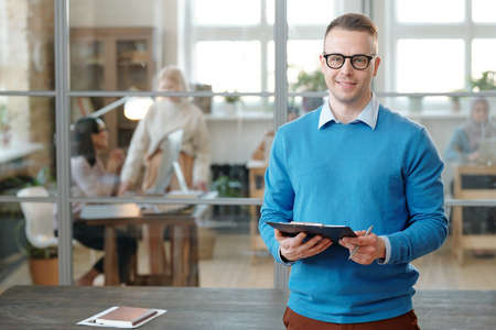 Portrait of smiling confident young Caucasian man in eyeglasses standing with clipboard against...