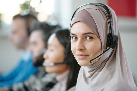 Portrait of smiling beautiful Arabian contact center agent in hijab using wireless headset with...